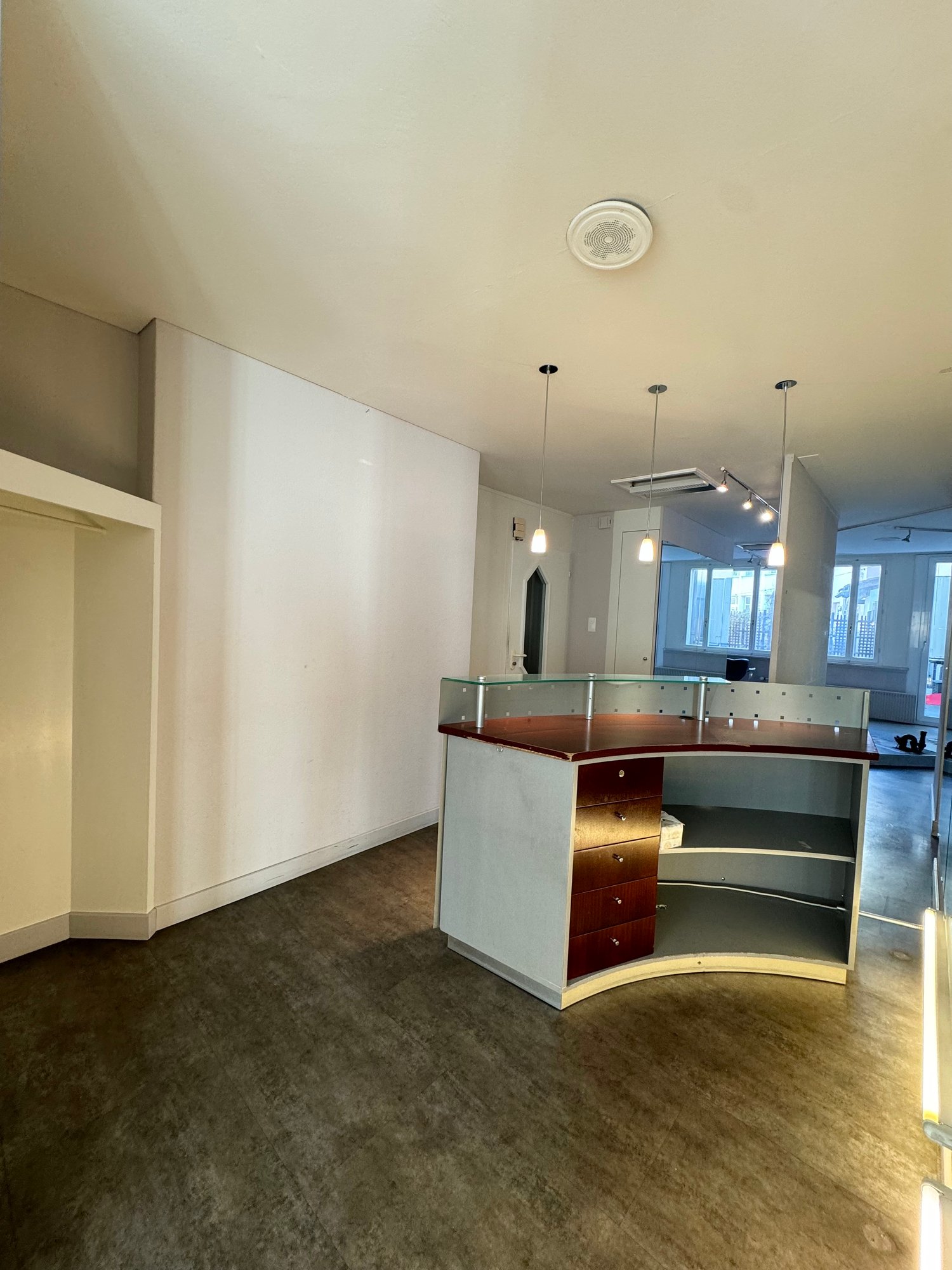 A room with an open layout, featuring a glass desk with wooden drawer and a glass top, and exposed white walls.