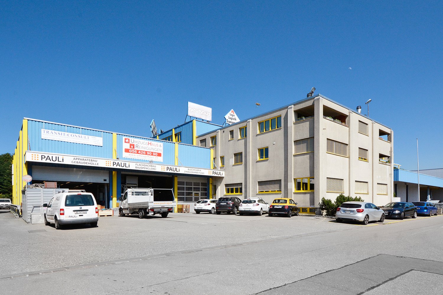 A multi-story commercial building with a Pauli logo and signage. The building has several garage doors and parking spaces in front. There are various vehicles parked in the lot.