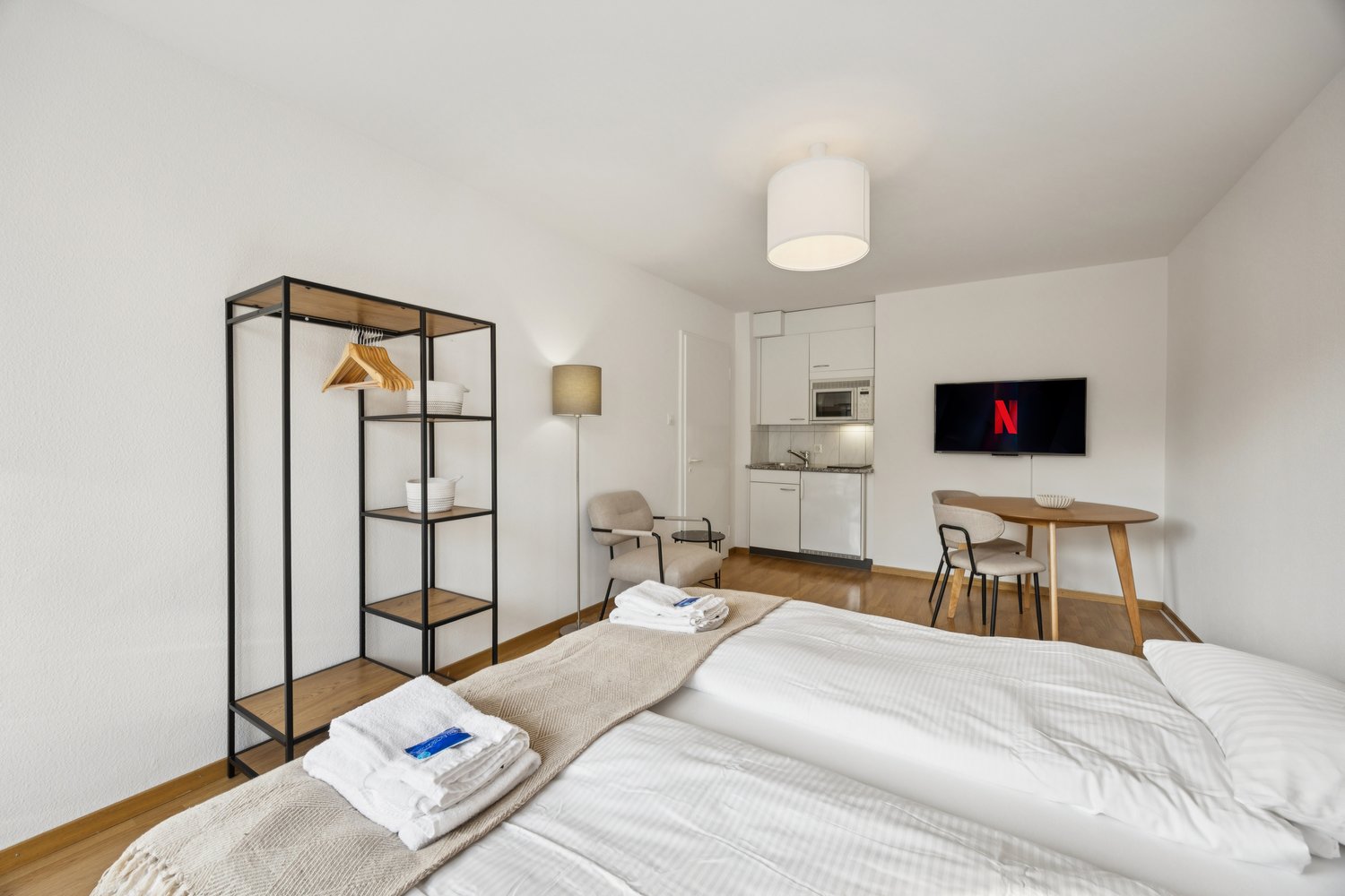 Compact bedroom with wooden floor, double bed, microwave, tv, dining table, small kitchen, hanging cabinet, floor lamp, and chair.