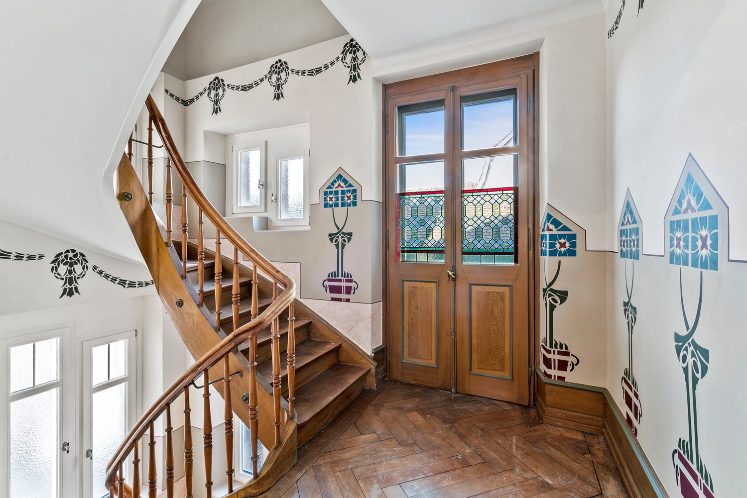 Wooden staircase with ornate railing, wooden double doors with stained glass windows, decorative wall art and patterns, hardwood flooring