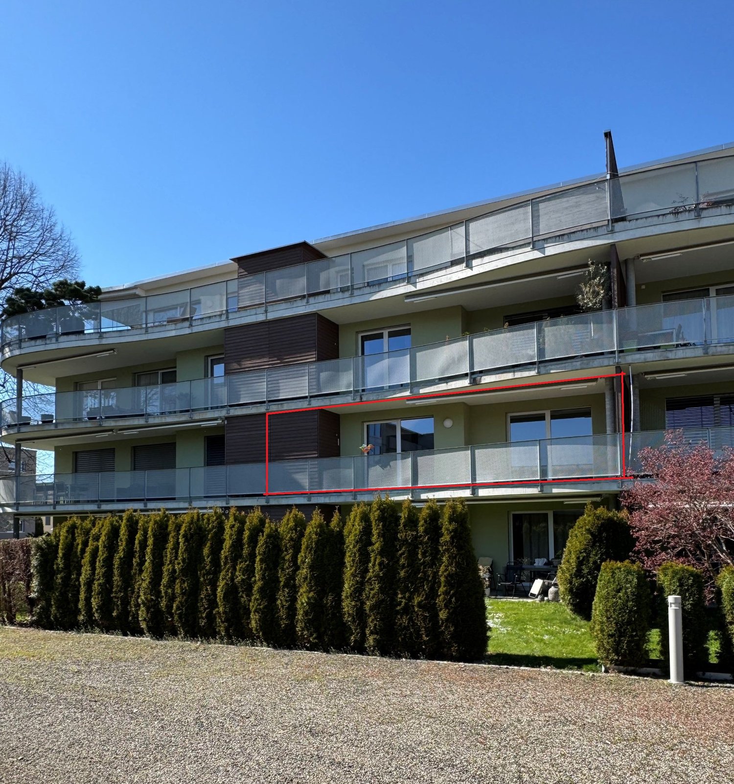 3 story apartment building, balconies, glass railings, green bushes, gravel driveway