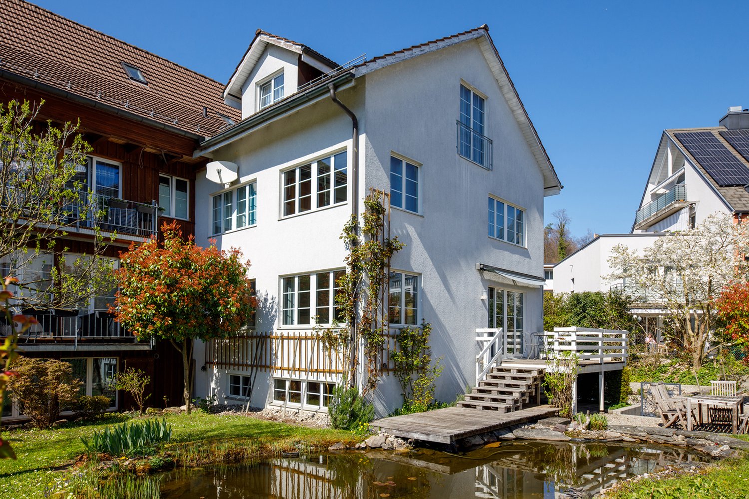 2 story house, white exterior, brown roof, large windows, wooden deck, small pond, garden