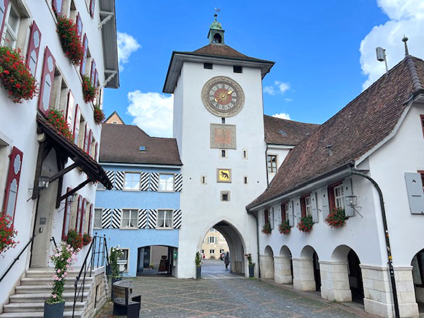 White building with clock tower, multiple windows with flower boxes, archway entrance, cobblestone street, archways along the building