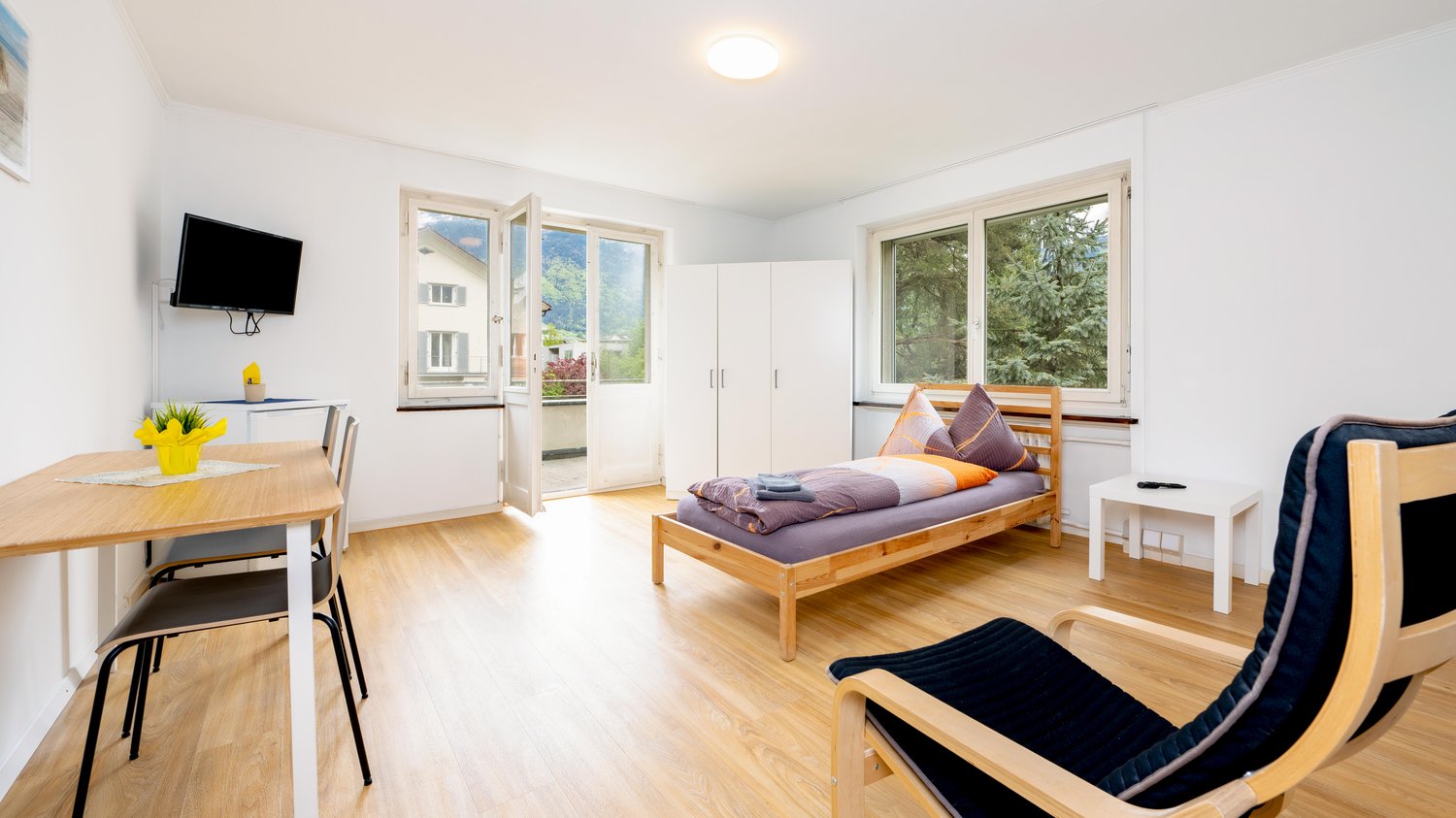 Bedroom: wooden bed, wooden desk with 3 chairs, side table, TV on wall, glass door leading to balcony, white wardrobe, round lamp