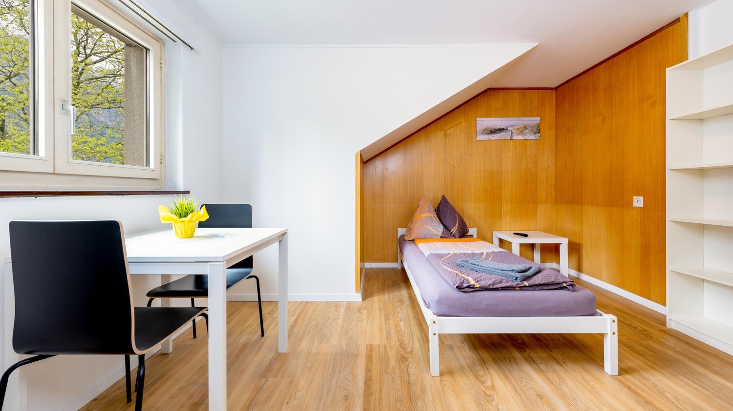 small room with a bed, desk, and chairs, wooden wall, window
