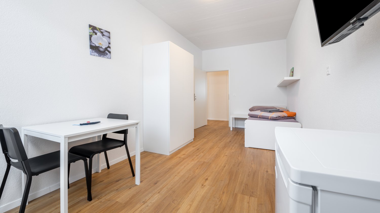 small room with a desk, two chairs, a bed, a wardrobe, a shelf, a television, and a washing machine