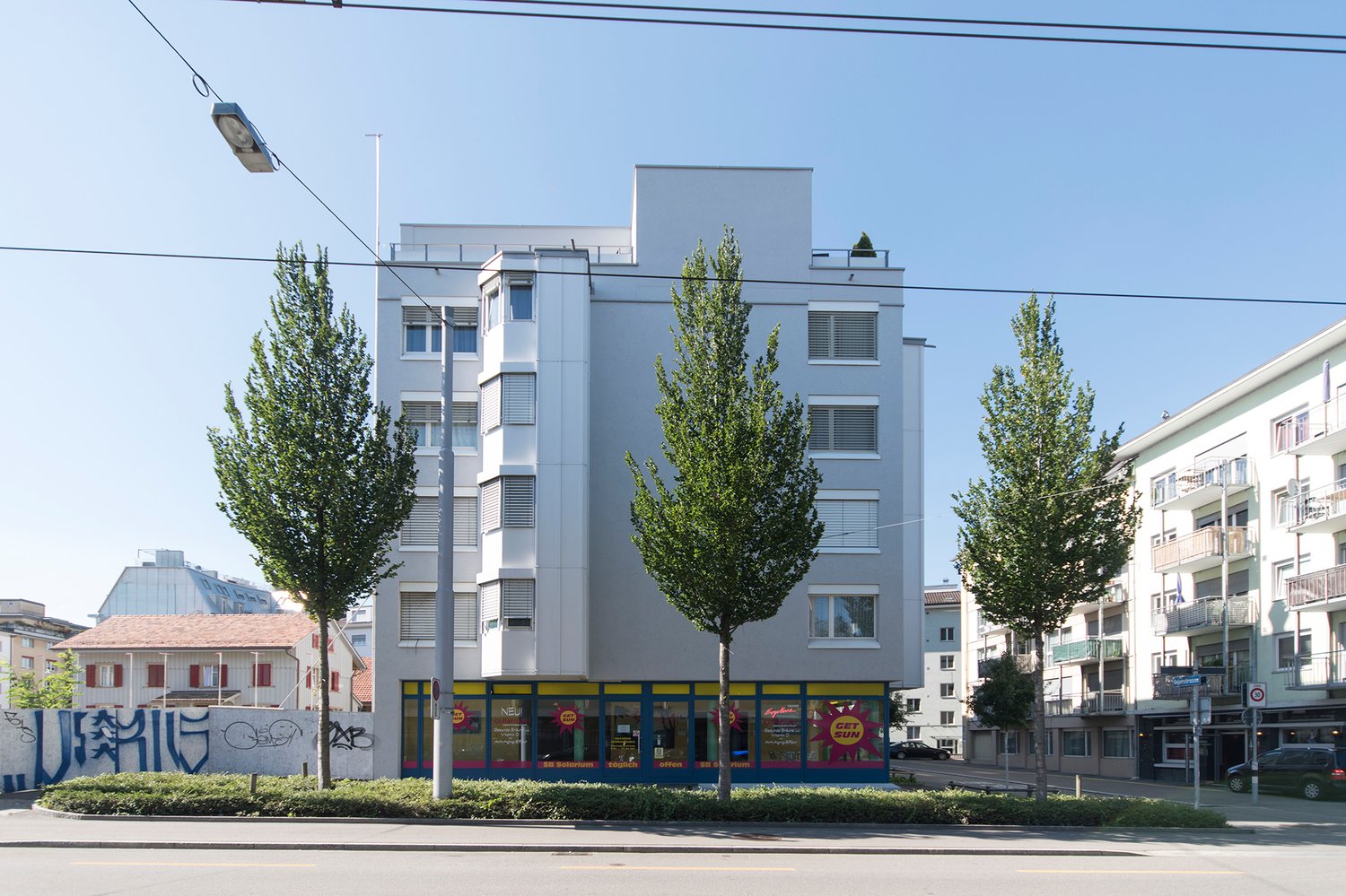 A multi-story commercial building with a modern, minimalist design. The building has large windows, balconies, and is surrounded by trees. There are several retail stores on the ground floor, including a Sun store.