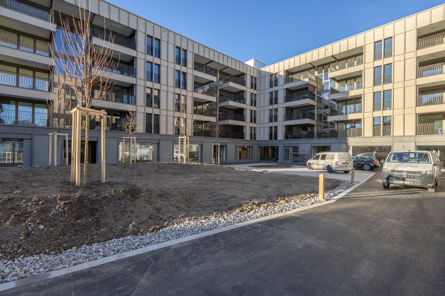 Multi-story apartment building with a modern, modular design. The building has balconies and large windows. There is a paved area with gravel and parking spaces in front of the building.