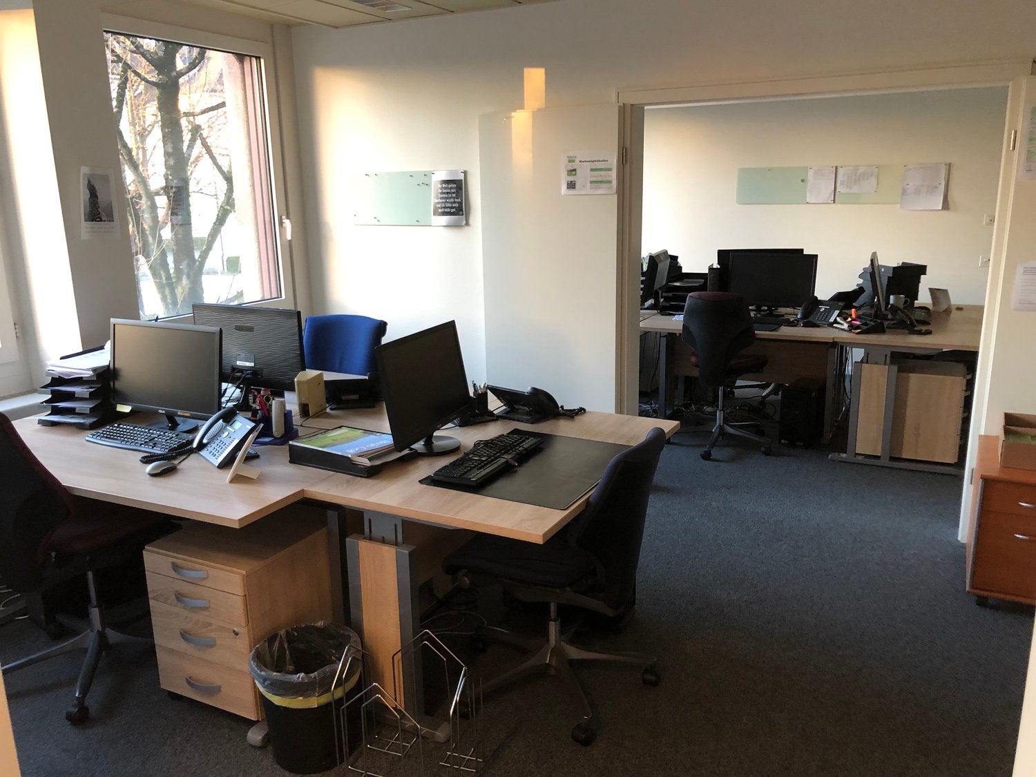 The image shows an office space with multiple desks, computers, and office equipment. The room has large windows providing natural light, and the walls are painted a light color. There are various office supplies and documents visible on the desks.