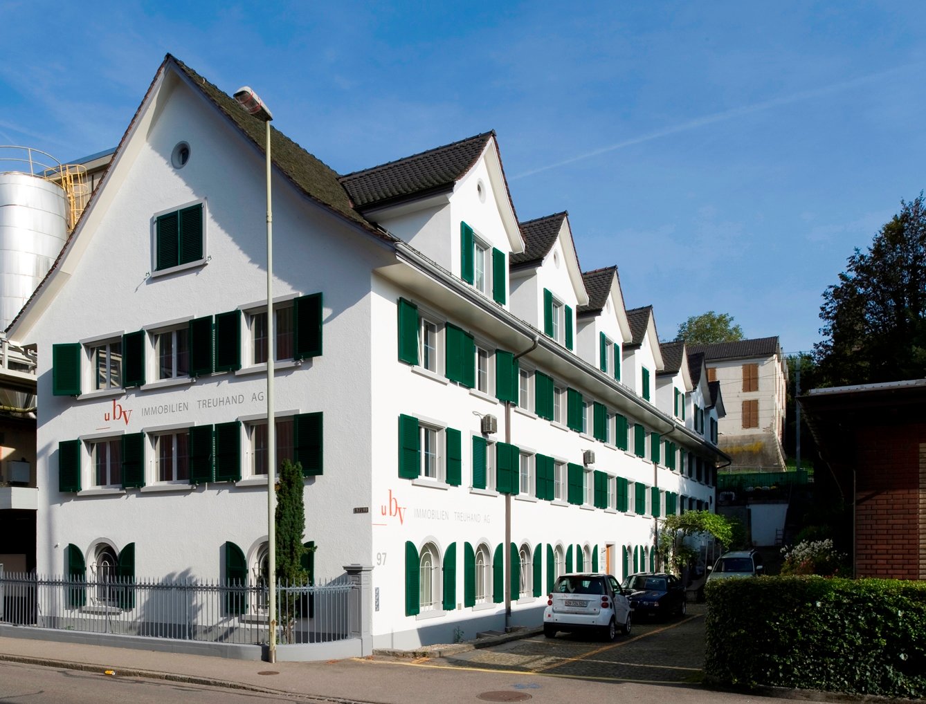 Modern white house with green shutters, gable roof, car parked outside, trees, brick building