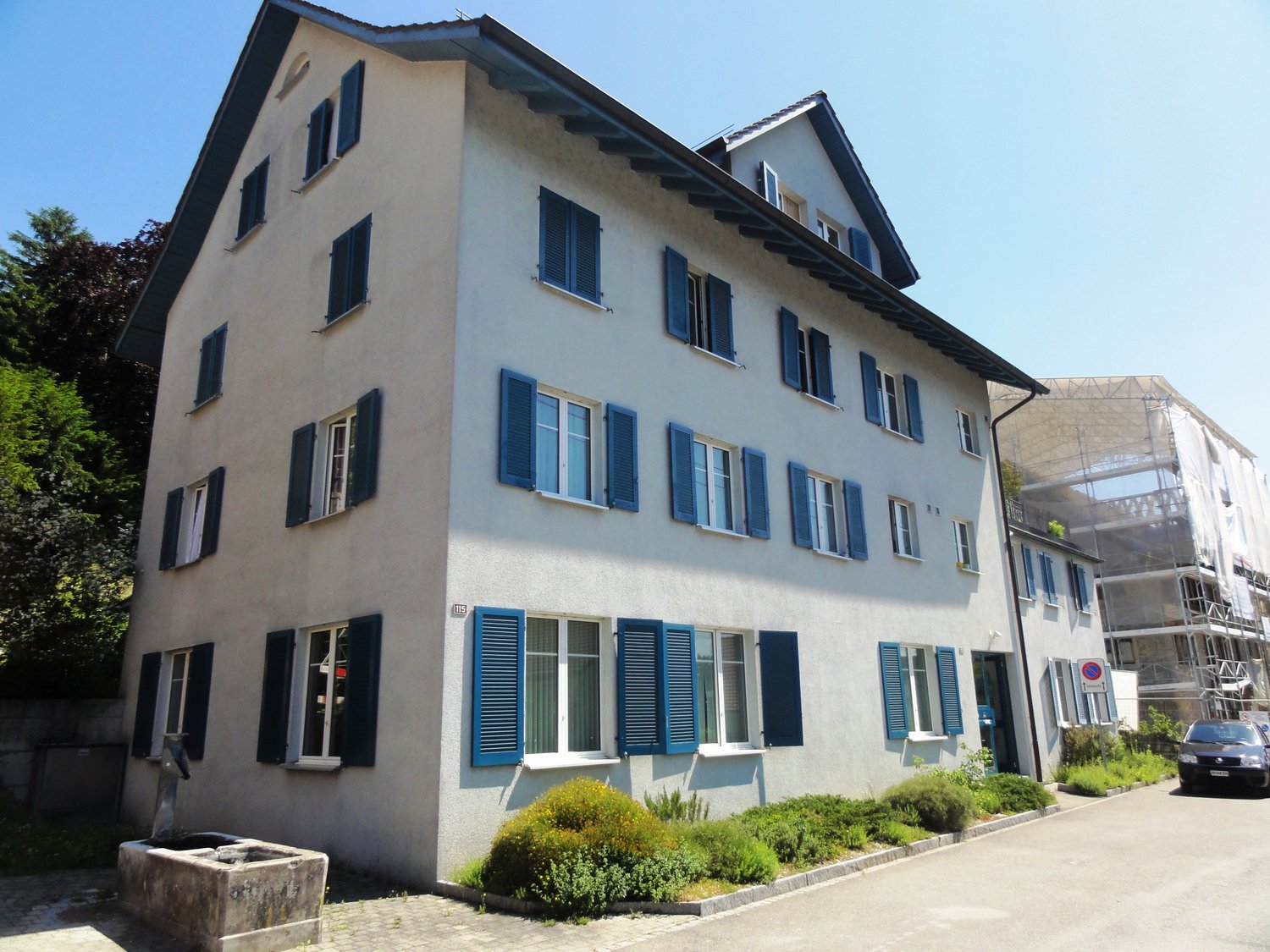 Apartment building, white walls, blue shutters, many windows, green plants, car parked on street