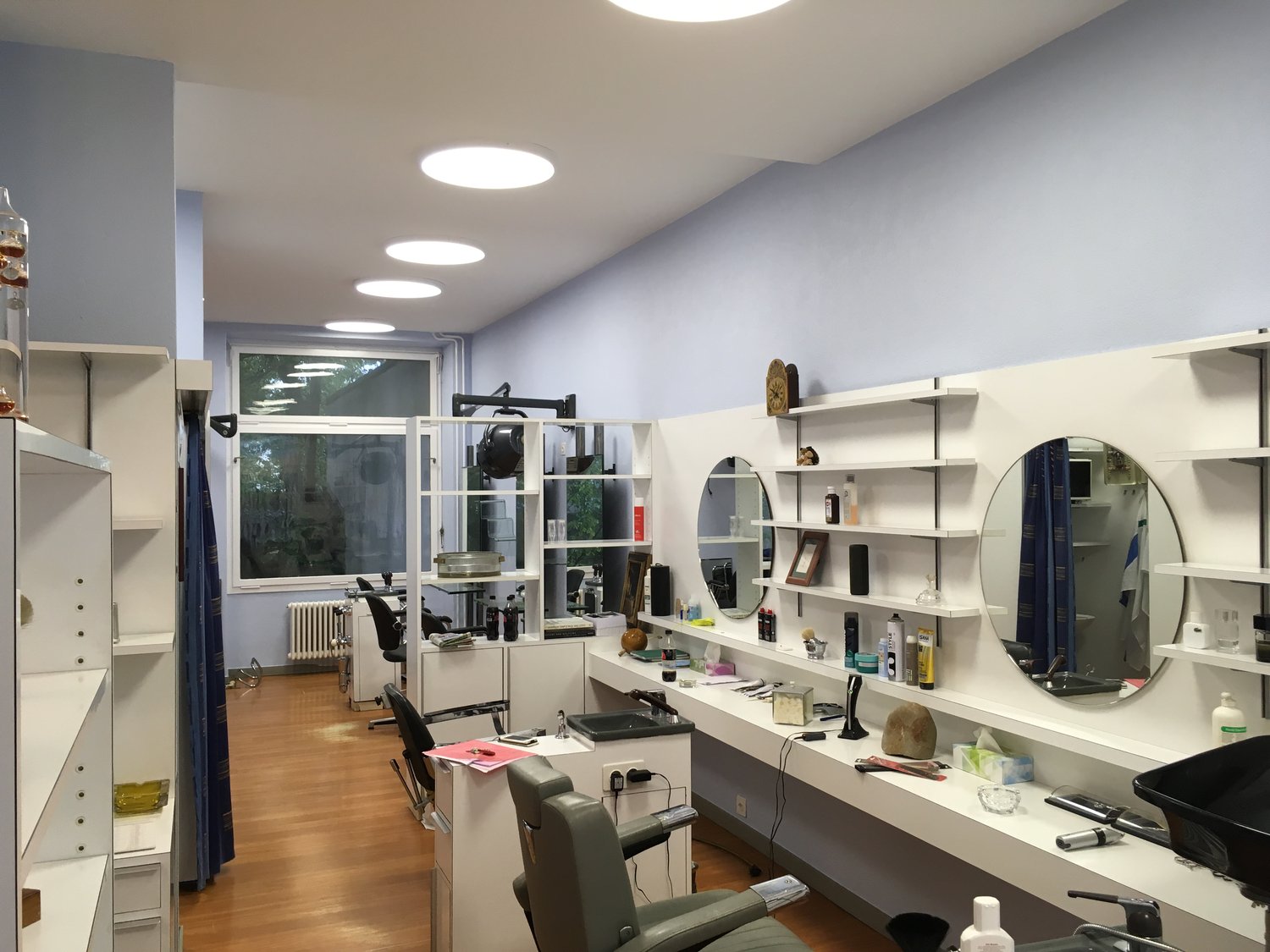 The image shows a room that appears to be a studio or office space. The room has gray walls, a large window, and various shelves and storage units along the walls. There are several mirrors and cosmetic items visible, suggesting it may be used as a beauty