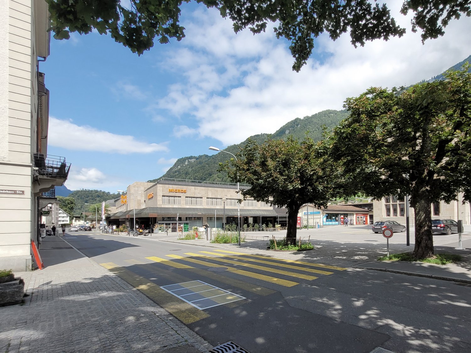 The image shows a street view of a town or city. There is a large supermarket building with the Migros logo visible. The street has a pedestrian crossing and parked cars. The surrounding area is lush with trees and greenery, and there are mountains visibl