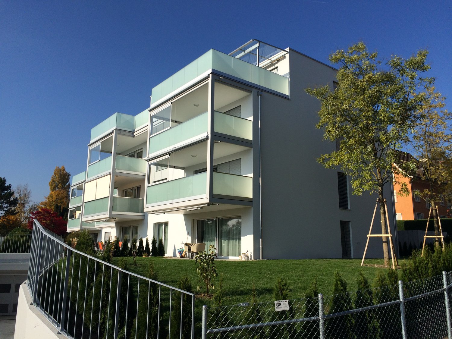 3 story apartment building with balconies, white exterior with glass railings, fenced yard, lawn, and trees