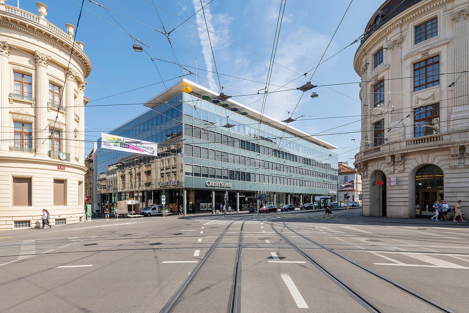 A large, modern glass and steel building with a distinctive architectural design, situated in a city center surrounded by historic, ornate buildings. The building appears to be a commercial or office space, with a Credit Suisse logo visible on the facade.