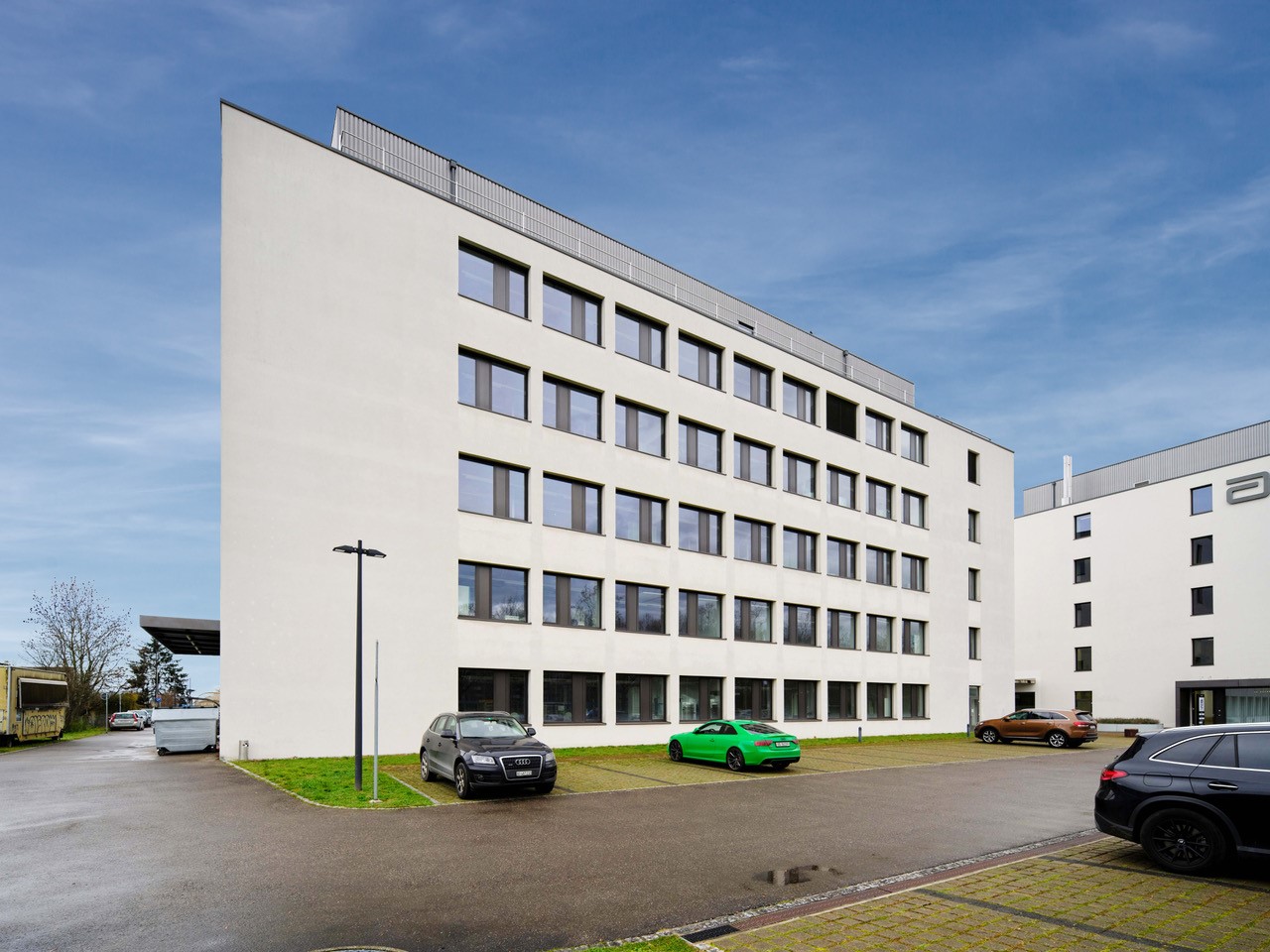 White modern office building, large windows, street lights, parking lot with multiple cars parked