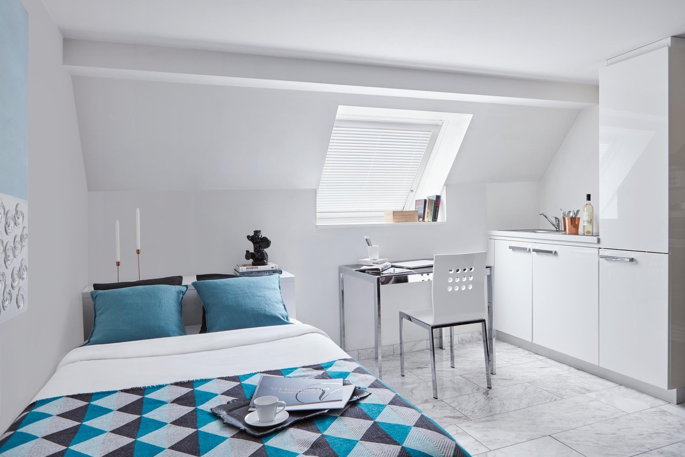 Single bedroom with bed, desk, chair, small kitchenette, bedspread, decorative items, window with skylight