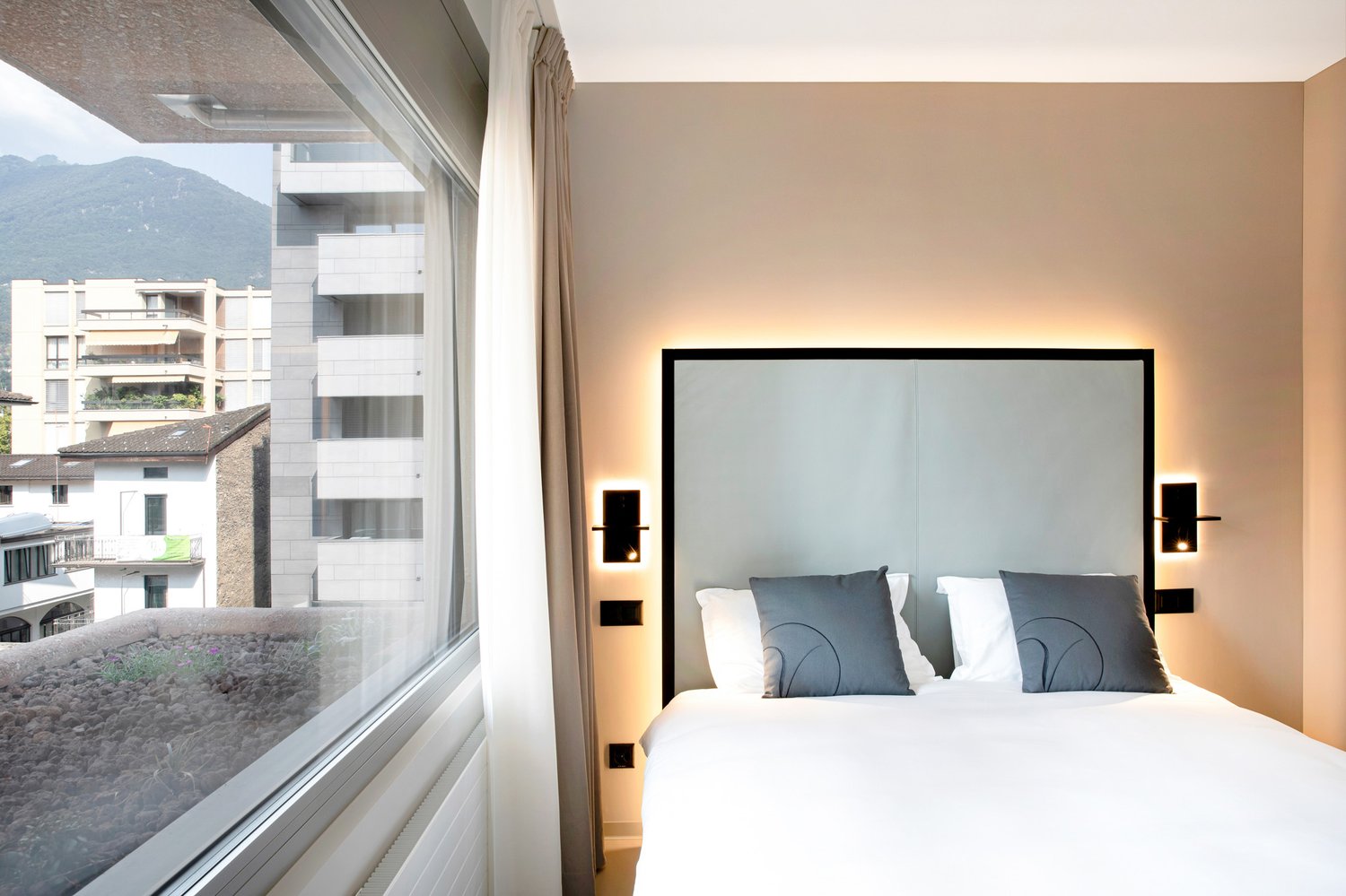 Double bed, large window with view of mountains, white walls, two bedside lights, gray headboard, white sheets, gray pillows
