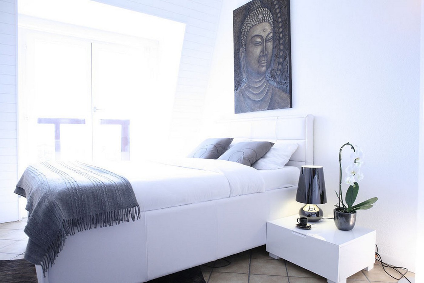 white bed, white nightstand, white lamp, white orchid, gray blanket, gray pillows, white walls, white ceiling, white floor, painting of Buddha