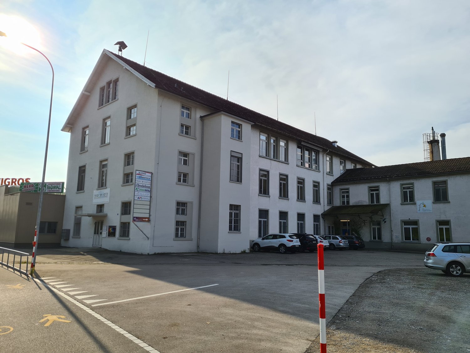 3 story building, white exterior, multiple windows, brown roof, parking lot, cars parked