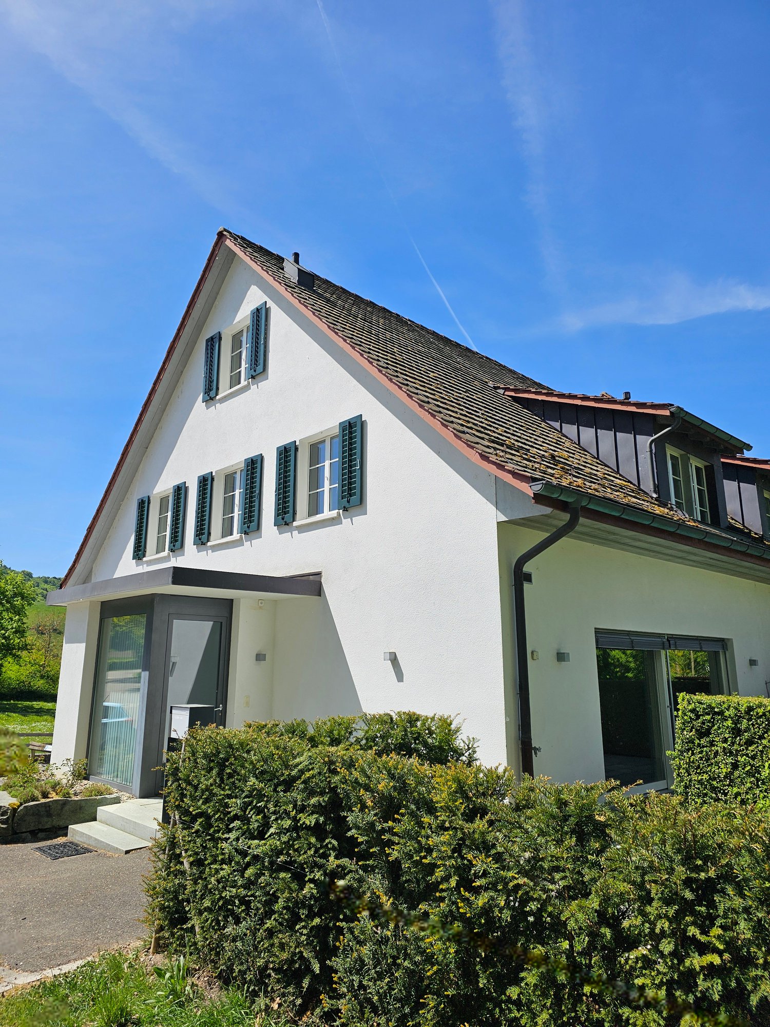 Modern white house with a brown roof, green shutters, and a glass door.