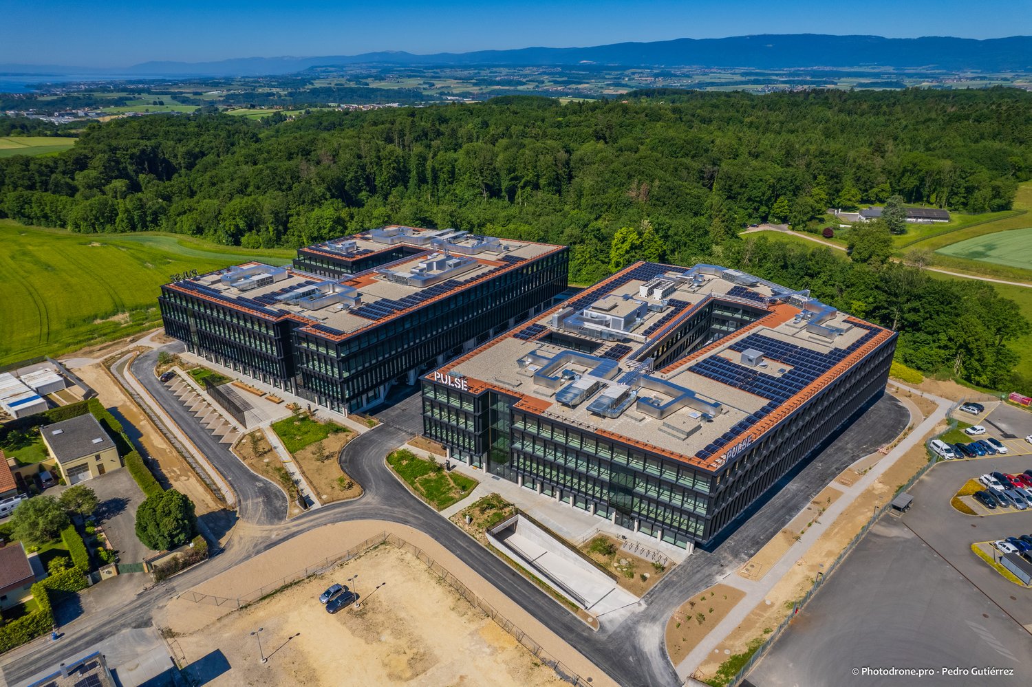 A large modern office complex with multiple interconnected buildings, featuring a sleek glass and steel design. The buildings have solar panels installed on the roofs, indicating a focus on sustainability. The complex is surrounded by lush green forests a
