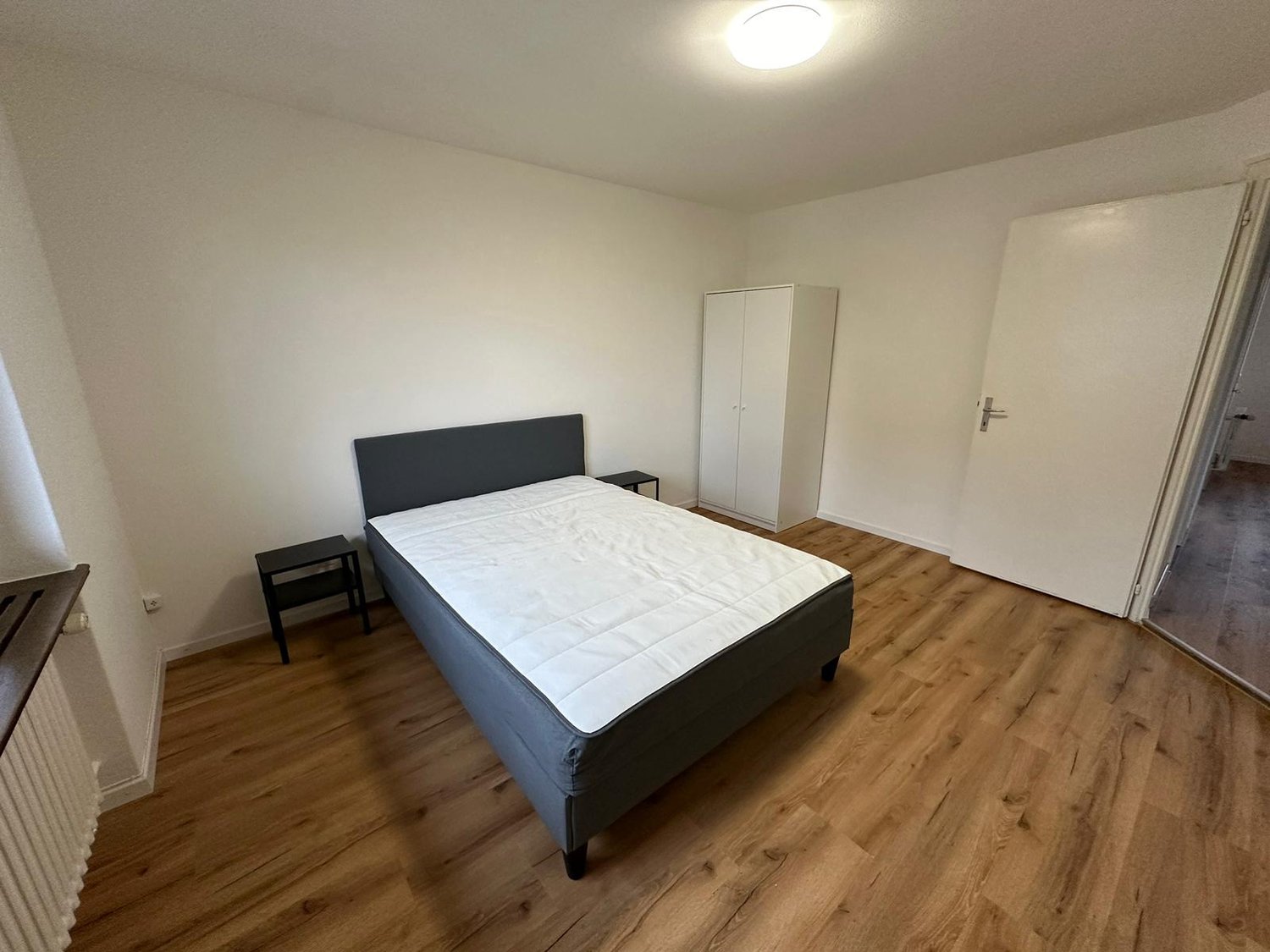 The image shows a bedroom with a double bed, a bedside table, and a wardrobe. The room has white walls and hardwood flooring.