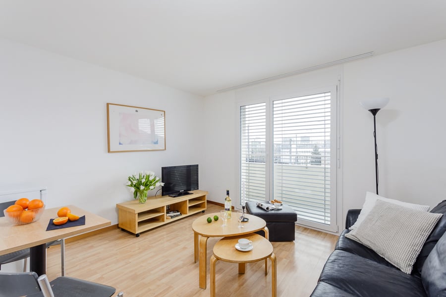 Bright and spacious living room with hardwood floors, white walls, large windows, a TV stand, coffee table, and a sofa.