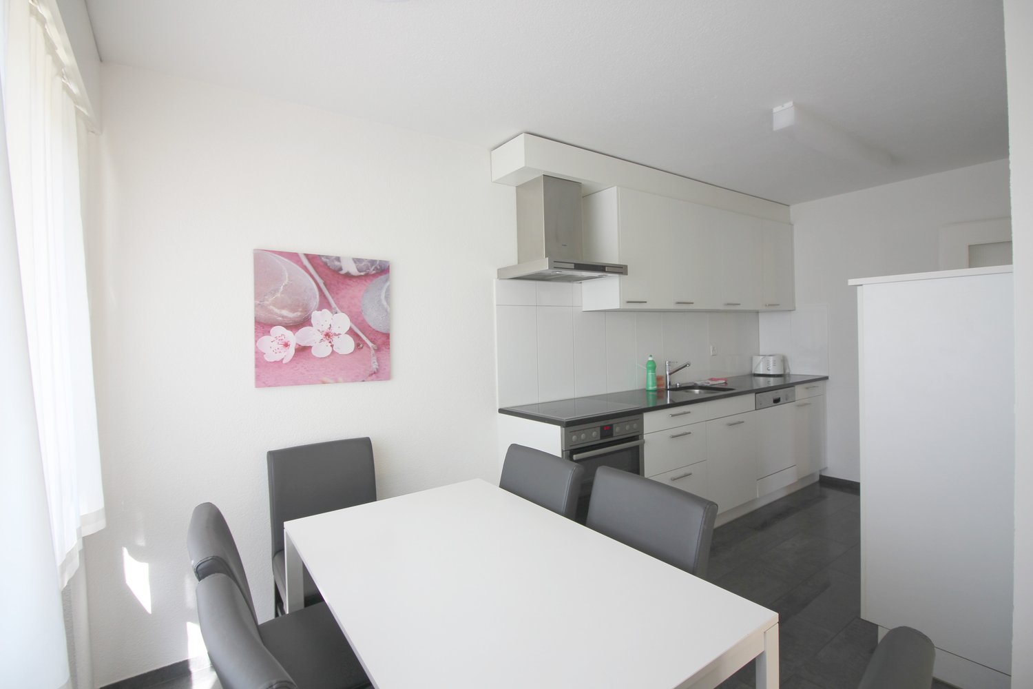 white tiled walls, kitchen with black countertop, white cabinets, modern appliances, grey chairs, dining table, artwork on wall, window with white curtains