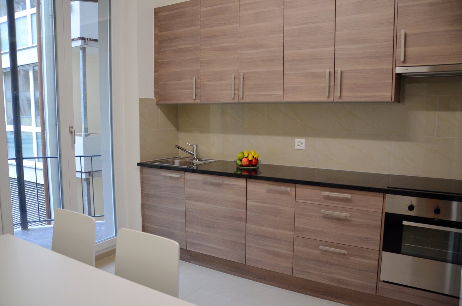 Modern kitchen with wood-paneled cabinets, black countertop, stainless steel sink, and a dishwasher. Large windows provide natural light and a view of the exterior. The kitchen also includes a dining area with white chairs.
