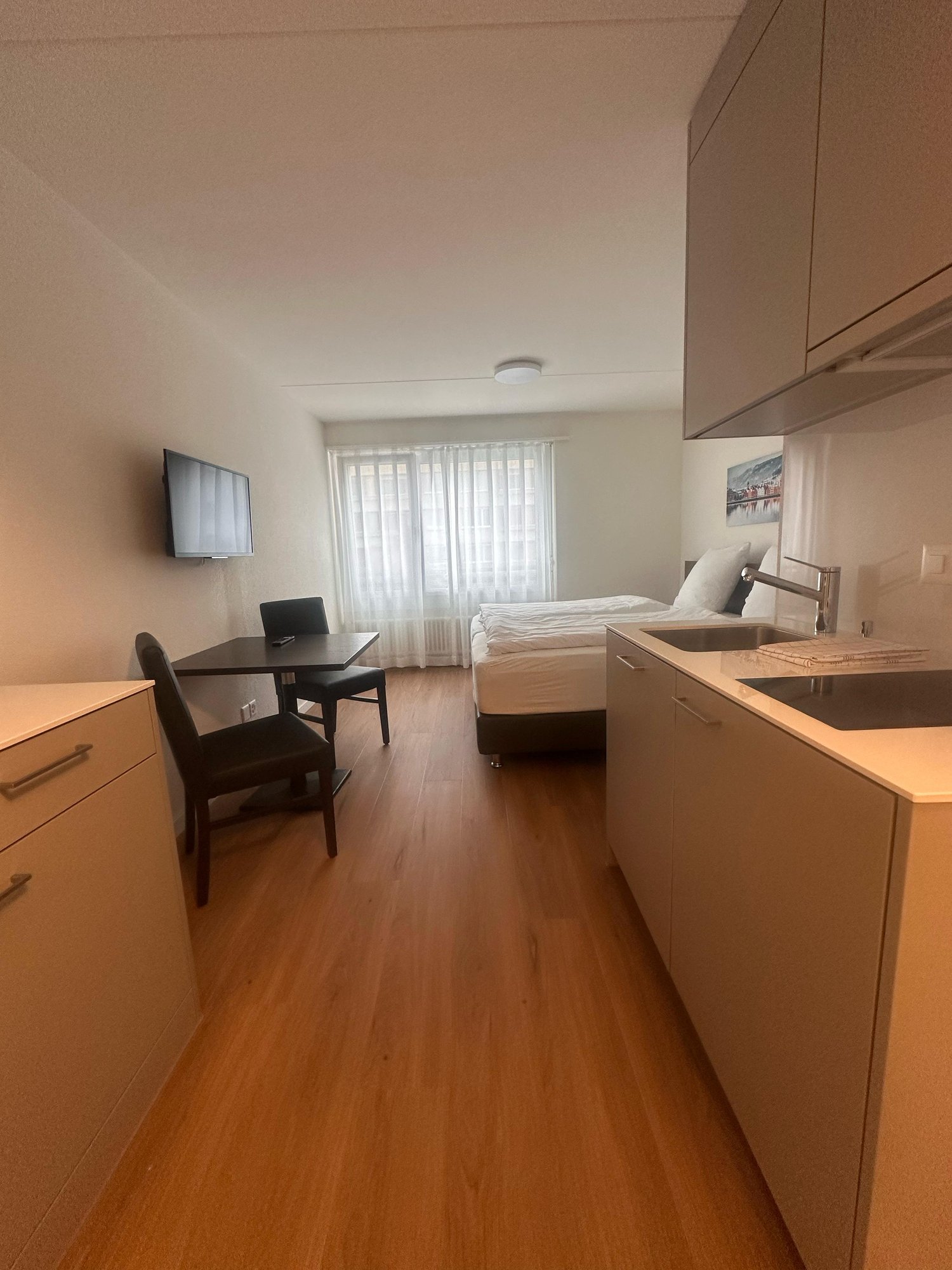Compact studio apartment with hardwood floors, white walls, a bed, a desk with a chair, and kitchen cabinets with a dishwasher visible.