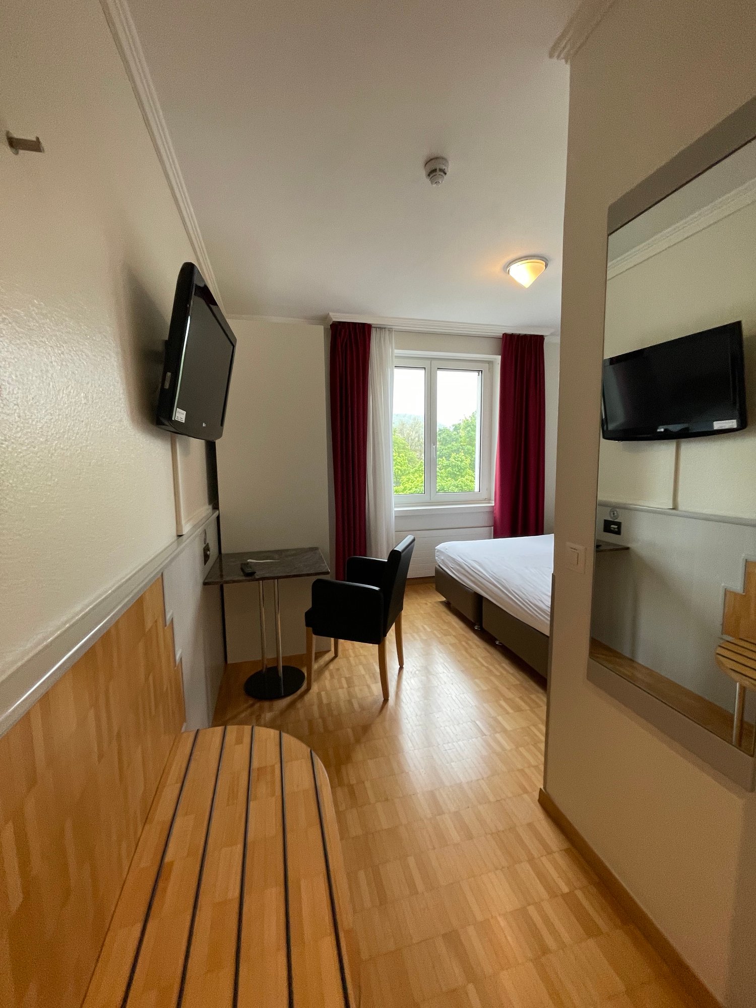 The image shows a bedroom with a bed, a desk, and two TVs mounted on the walls. The room has hardwood floors and large windows with red curtains. The overall decor appears to be modern and minimalist.