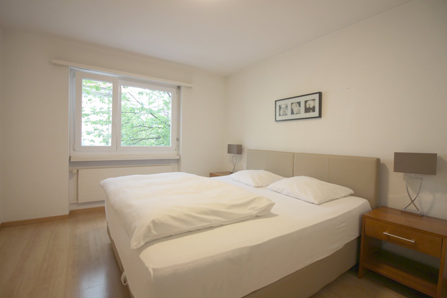 Spacious bedroom with large windows overlooking greenery, white walls, hardwood floors, a king-size bed with white bedding, a wooden nightstand, and a framed artwork on the wall.