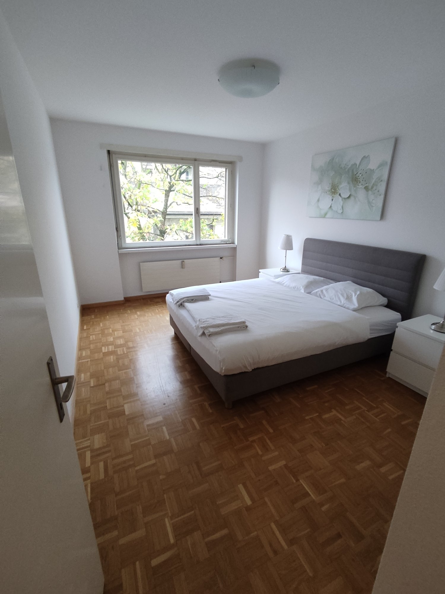 Spacious bedroom with large window overlooking trees, hardwood parquet flooring, grey upholstered bed frame, and a decorative floral painting on the wall.
