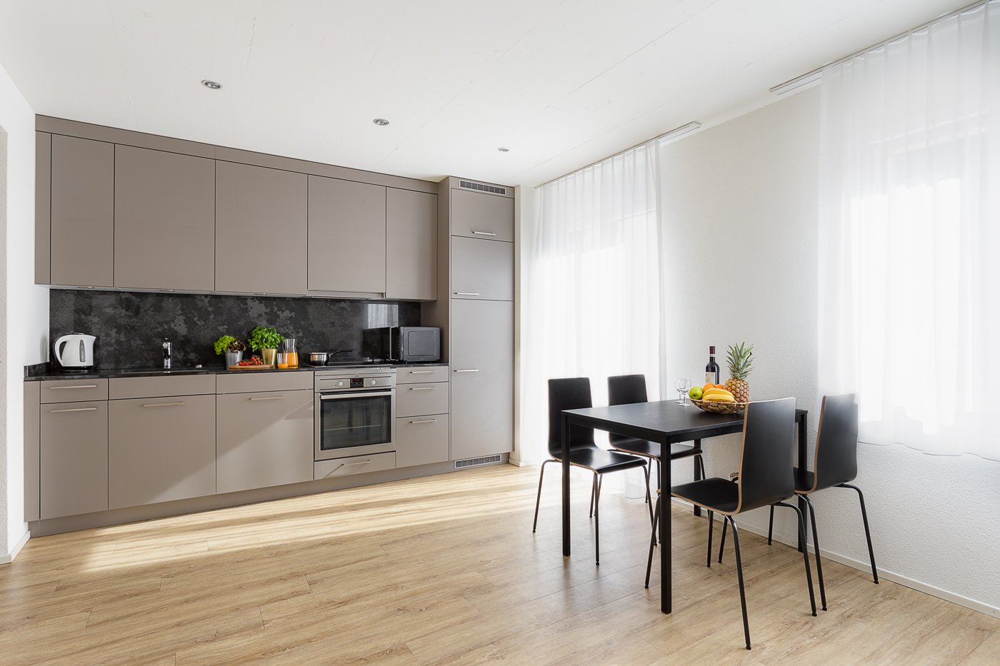 Modern kitchen with wood flooring, gray cabinets, oven, wooden dining table with chairs, bowl of fruit, bottle, glasses