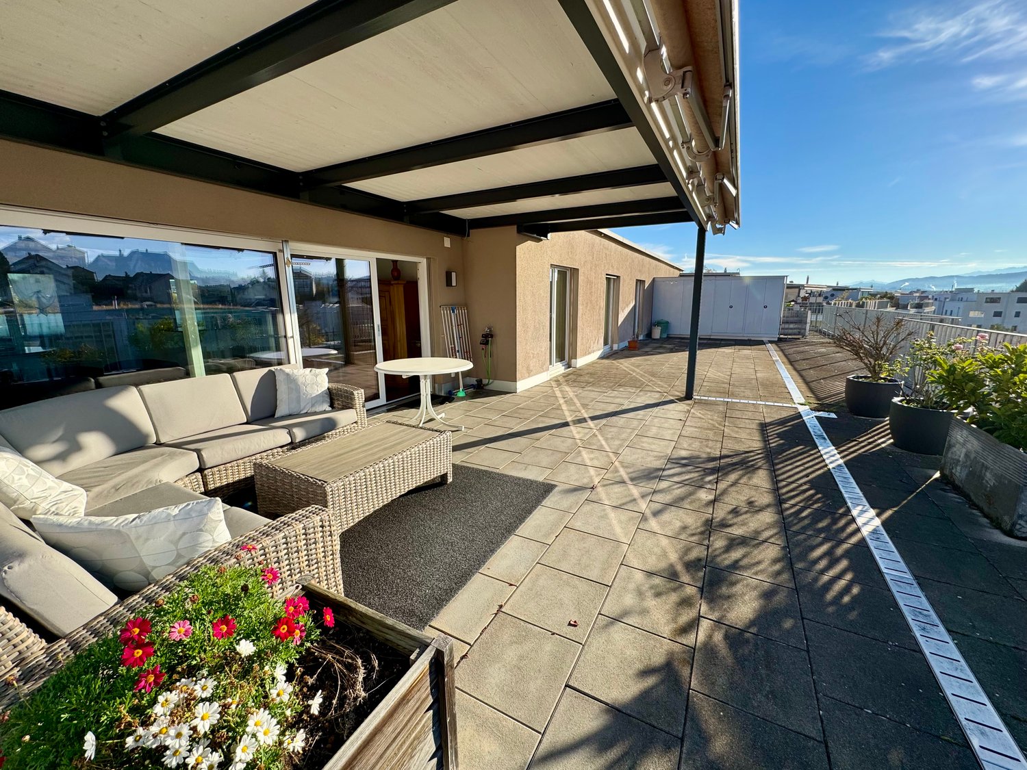 A covered outdoor terrace with a view, featuring a patio furniture set, a table, a large flower pot with flowers, and a storage box on the right side.