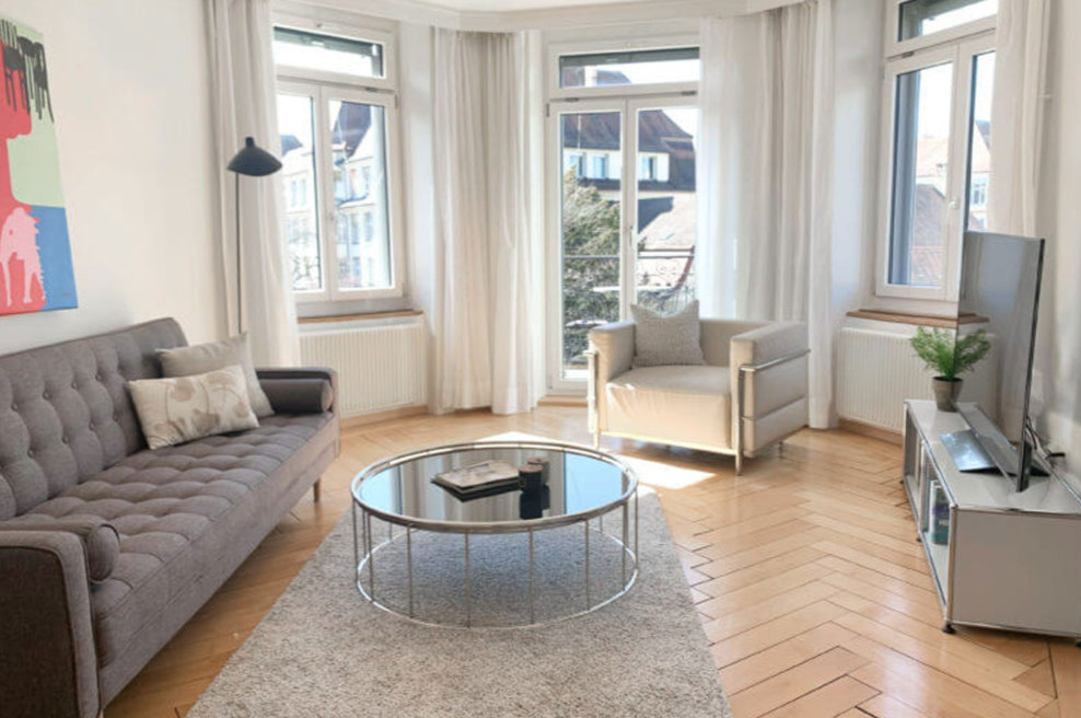 living room, grey sofa, circular glass coffee table, armchair, floor lamp, white curtains, radiator, white walls, parquet flooring, windows, flatscreen TV, flat screen TV