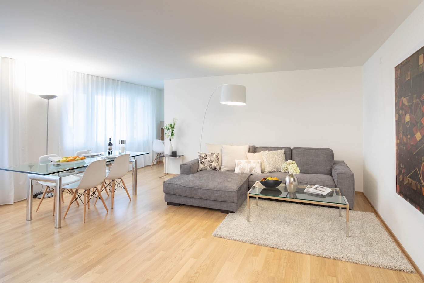 modern living room with glass table, comfortable gray couch, glass coffee table, rug, floor lamp, white walls, wooden floor