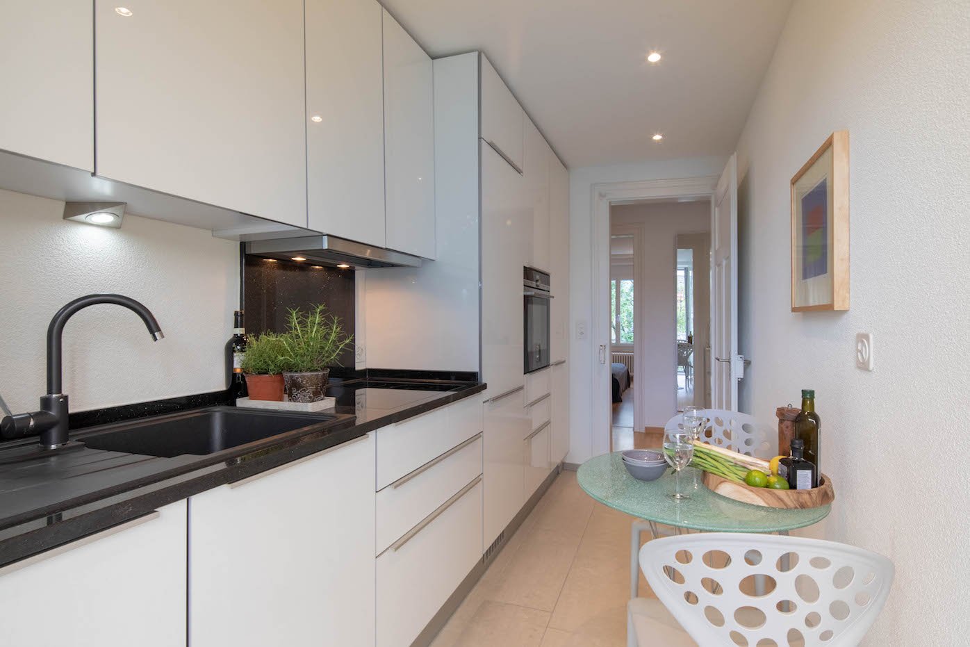 Kitchen: white cabinets, black countertops, modern faucet, integrated oven, wine bottles, fresh produce, dining table, white chairs, artwork on wall