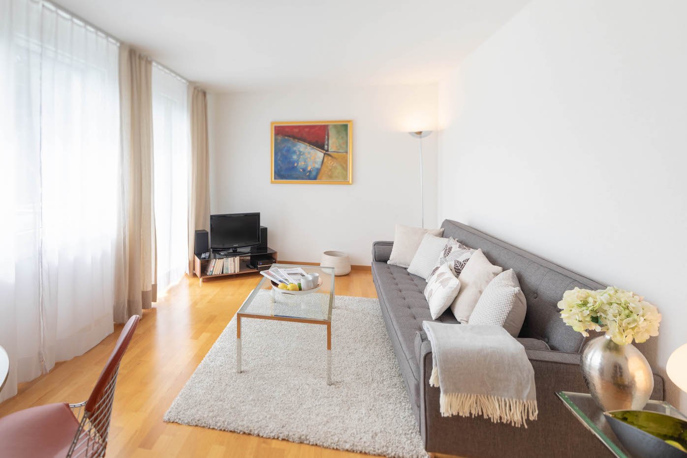 Wooden floor, large sofa, table with flowers, TV, white walls, glass table