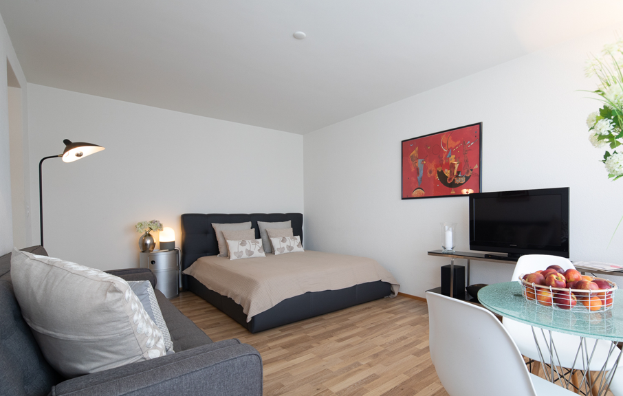 small room with a bed, couch, lamp, tv, dining table with fruit, nightstand with lamp and picture