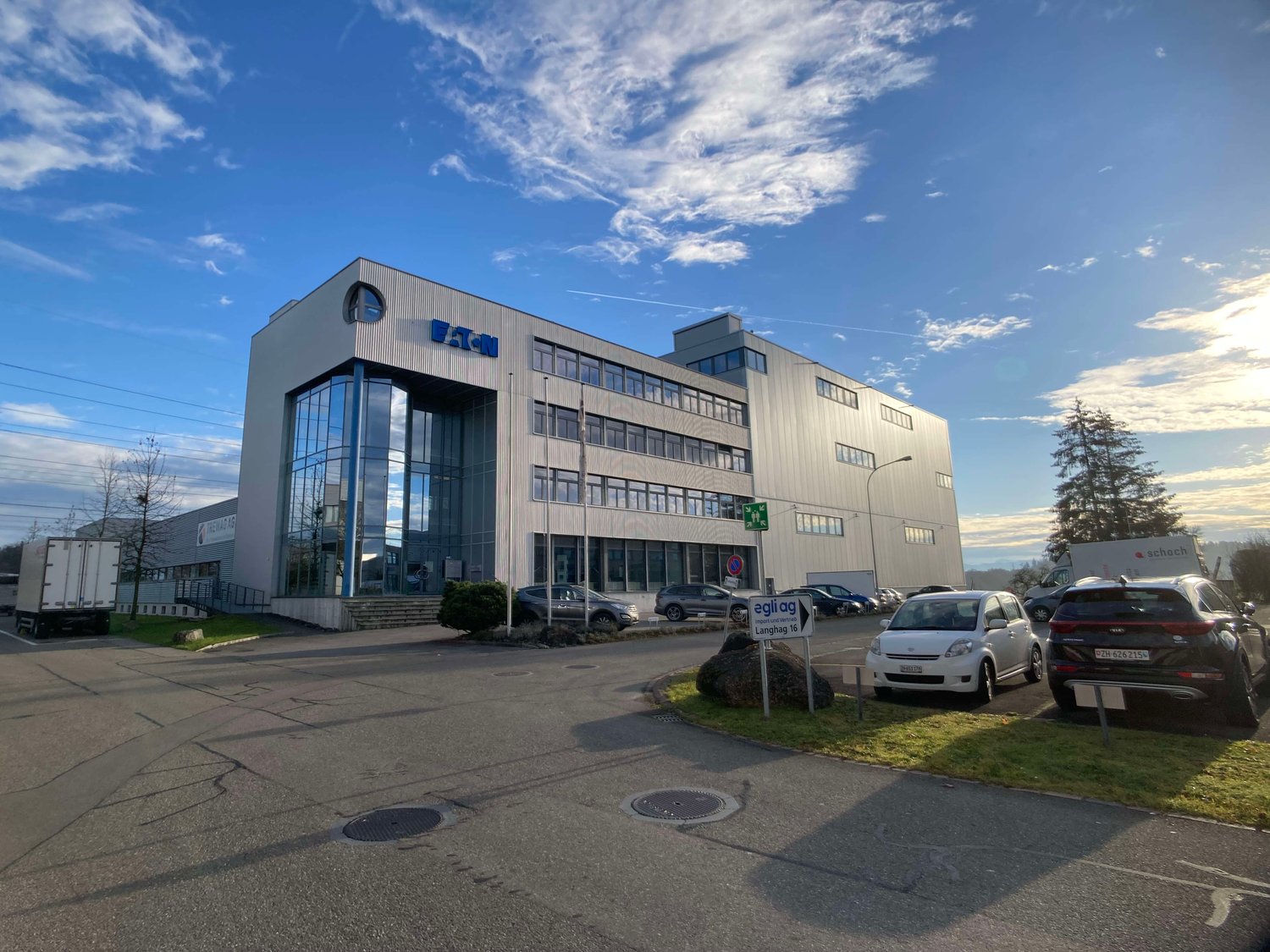 A multi-story office building with a modern, industrial-style architecture. The building has a large glass facade and is surrounded by a parking lot with several cars parked. The sky is partly cloudy with a blue background.