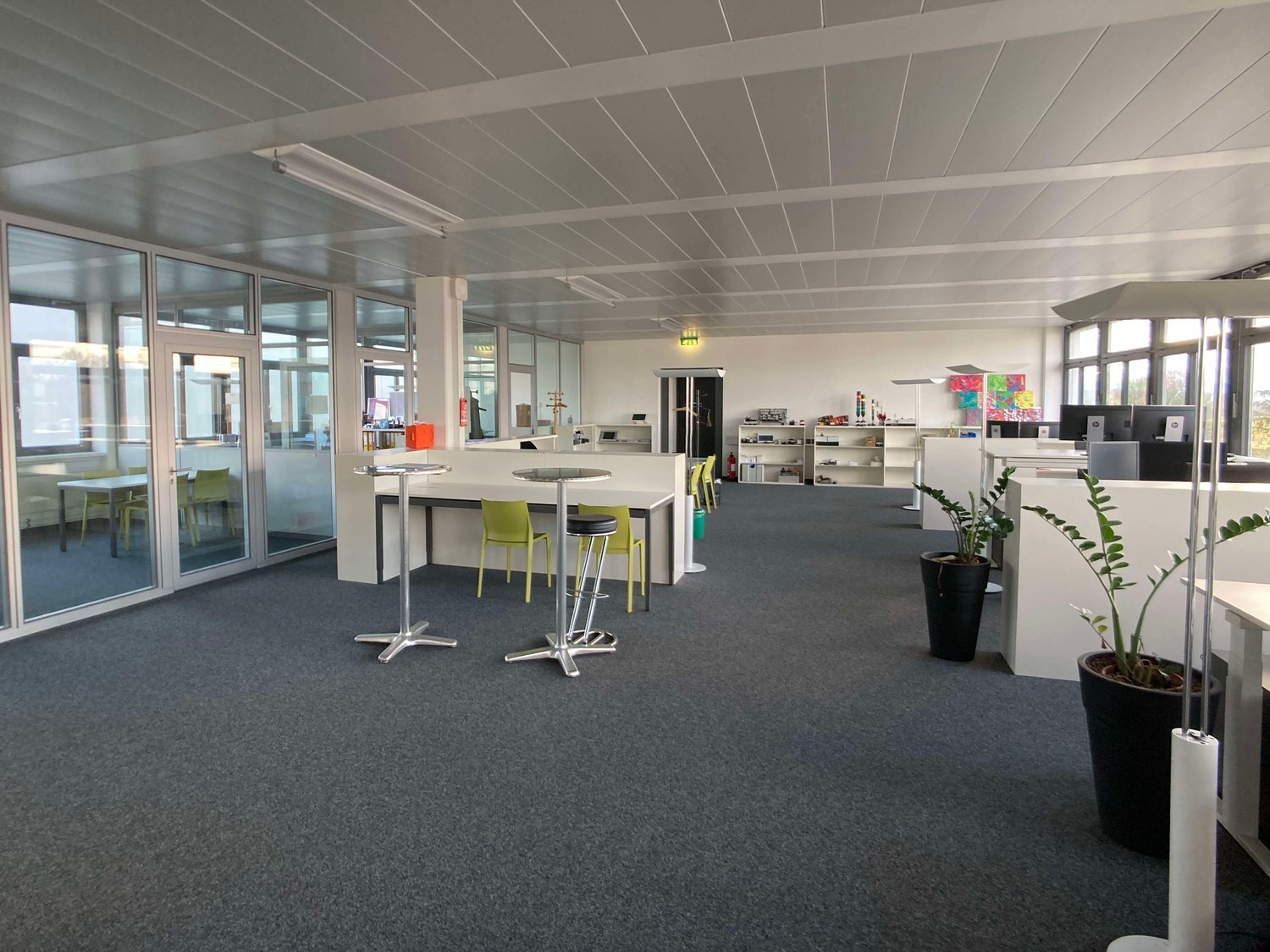 The image shows a modern and open office space with a high ceiling, glass walls, and a mix of workstations and lounge areas. The space has a gray carpet floor, white walls, and a gray ceiling with exposed beams. There are several desks with chairs, shelve