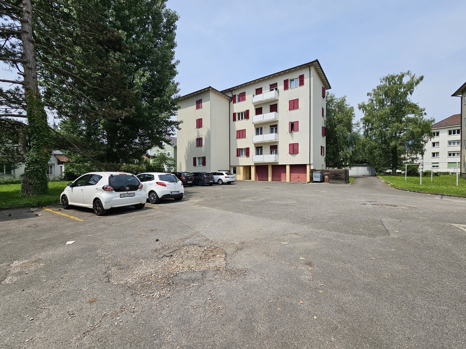 4-story apartment building with white exterior and red shutters, parking lot in front of the building, surrounded by trees