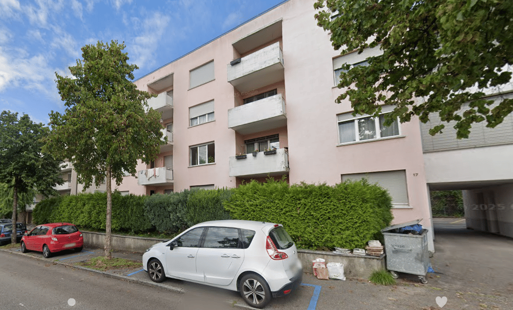 3-story apartment building with balconies, surrounded by trees and greenery, with parked cars in the foreground