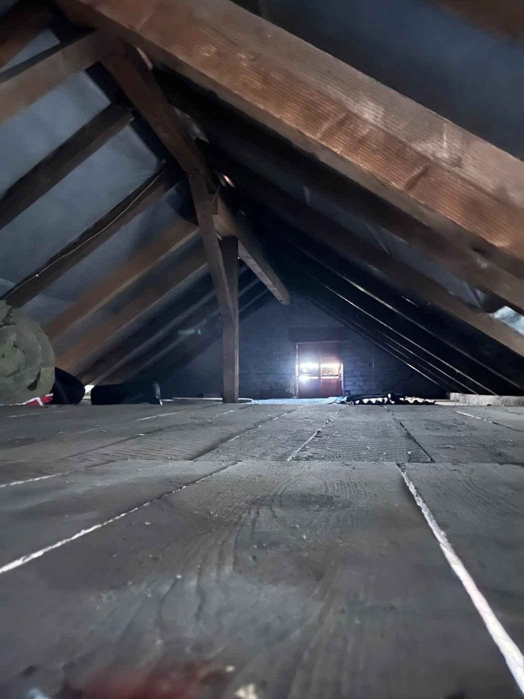 The image shows the interior of an attic space. The wooden beams and trusses are visible, along with a small window that provides some illumination. The floor appears to be made of wooden planks or boards. The overall atmosphere is dark and shadowy, sugge
