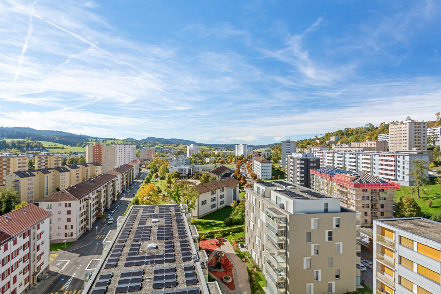 Aerial view of an urban setting with residential buildings, rooftop solar panels, greenery, and distant mountains under a clear sky
