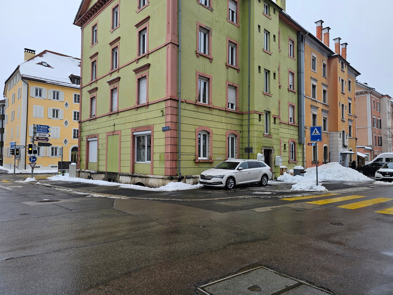 Corner building with four floors, light green color with red frames around the windows, snow on the ground, a parked car in front, two other buildings next to it