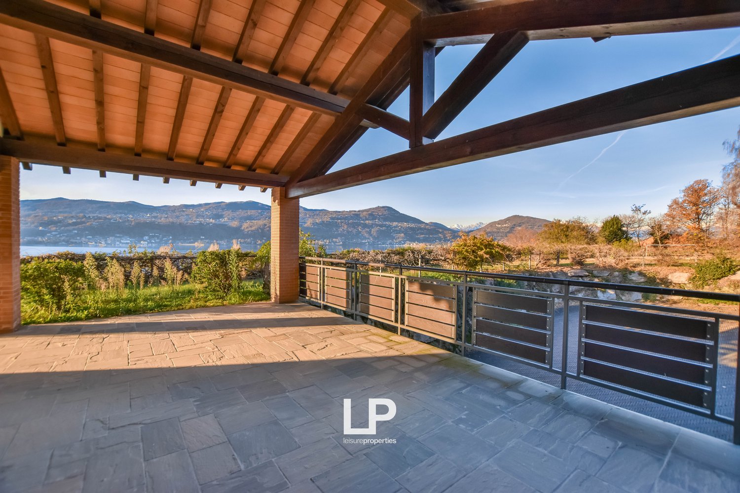 Covered outdoor terrace with wooden beams and tiled flooring, offering a scenic view of the surrounding mountains and lake in the distance.