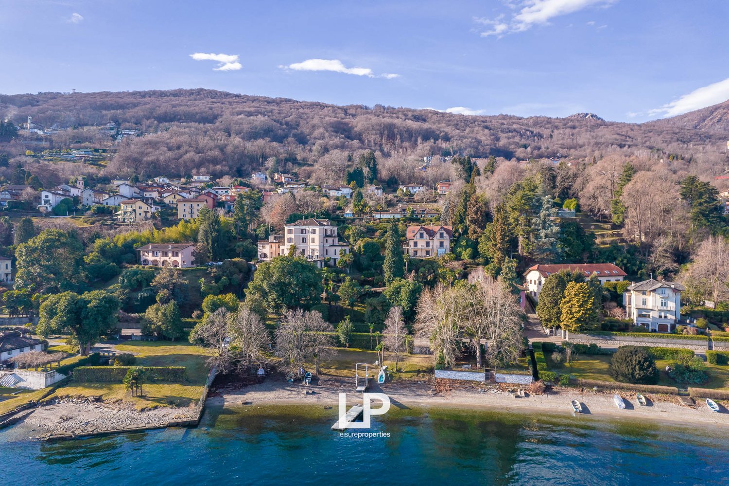 Aerial view of a hillside town with several houses, surrounded by trees and mountains, with a shoreline and boats